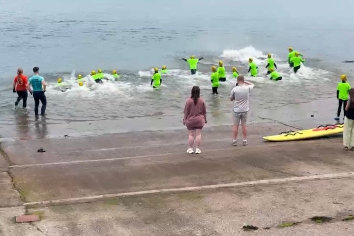 Group in neon green shirts on beach, some entering water; three people and a paddleboard onshore.