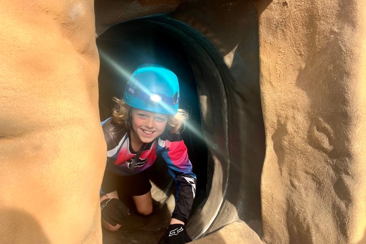 Person wearing helmet crawls out of a rock tunnel.