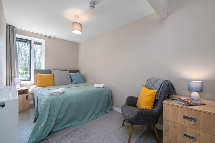 Cozy bedroom with a bed, colorful pillows, armchair, nightstands, and a window with natural light.