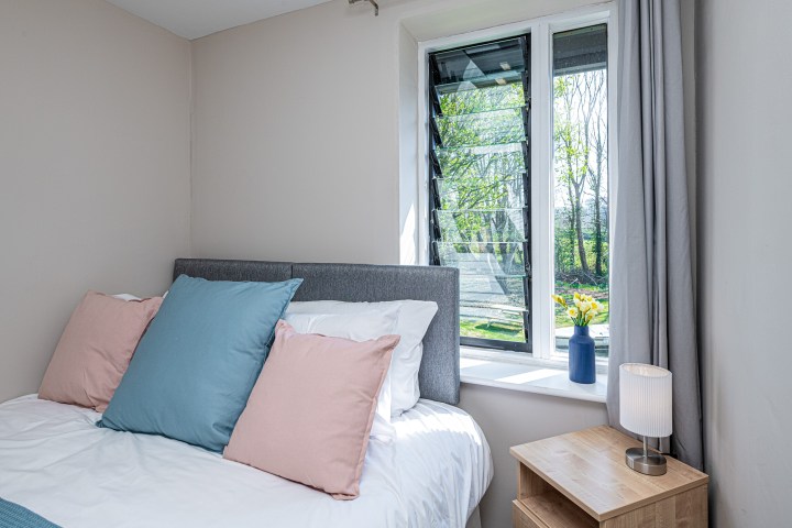 Bedroom with a bed, colorful pillows, a window, and a nightstand with a lamp and flowers in a vase.