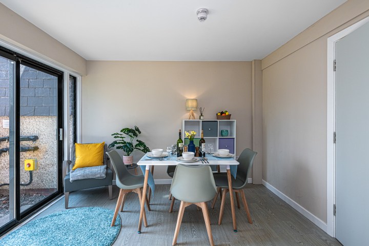 Modern dining room with table set for four, armchair, plant, and shelving unit under soft lighting.