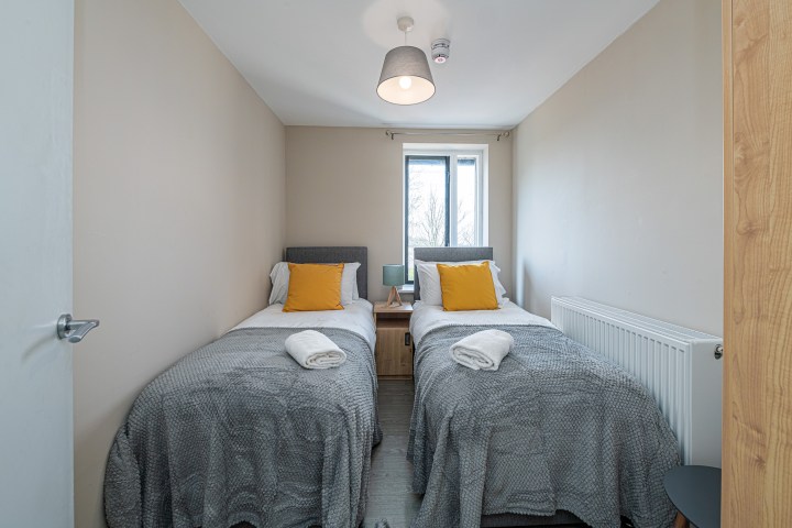 Two single beds with gray blankets and yellow pillows, in a room with a window and a small lamp.