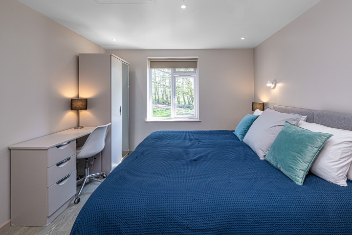 Bedroom with double bed, blue bedding, desk with chair, and window view of trees.