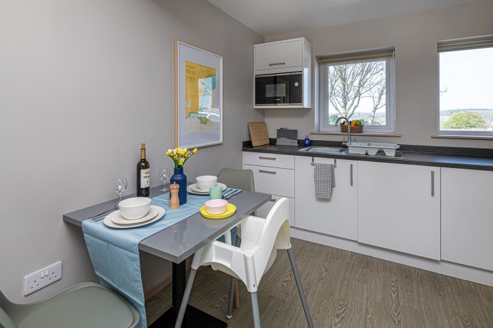 Modern kitchen with table set for two, white cabinets, and a window view of trees.