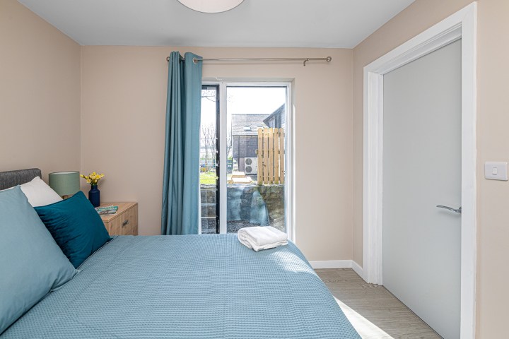 Bedroom with blue bedspread, window with blue curtains, dresser with lamp and flowers.