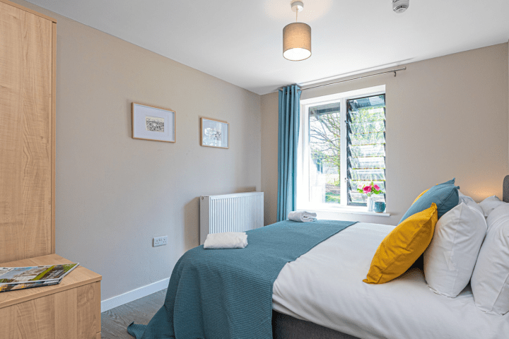 Cozy bedroom with blue and yellow cushions, window, and artwork on the walls. Wooden cabinet with magazines.