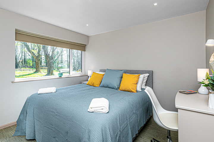 Cozy bedroom with blue bedspread, yellow pillows, desk, chair, flowers, and large window overlooking trees.