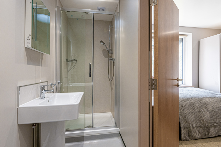 Modern bathroom with shower, sink, and mirror adjacent to a bedroom with a bed and wardrobe.