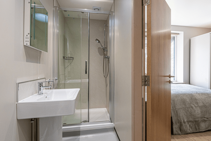 Modern bathroom with shower, sink, and mirror, adjacent to a bedroom with a bed and window.