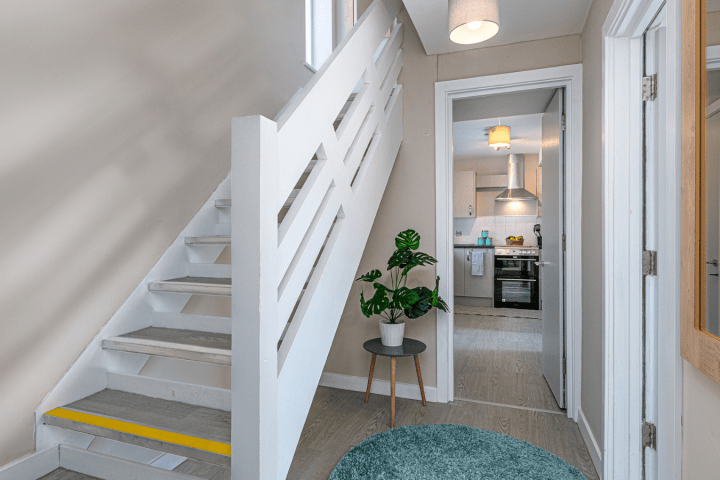Hallway with stairs, plant on table, mirror, and view into a kitchen.