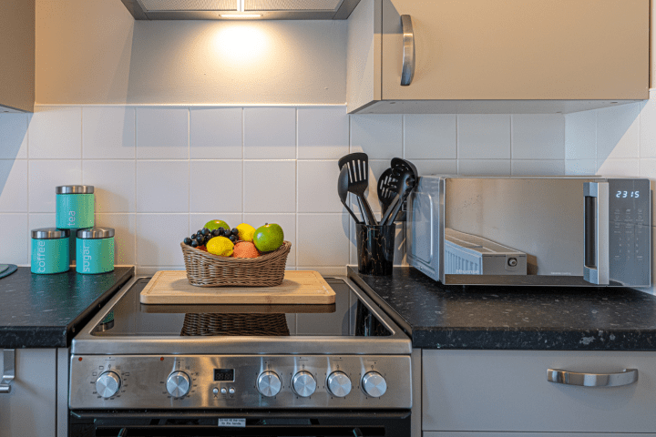 Modern kitchen with stove, fruit basket, utensils, and microwave on black countertops.