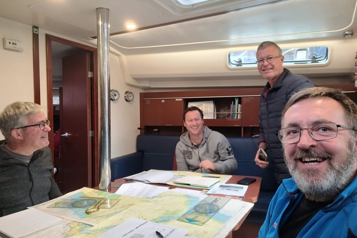 Four people smiling around a table with nautical charts on a boat cabin.