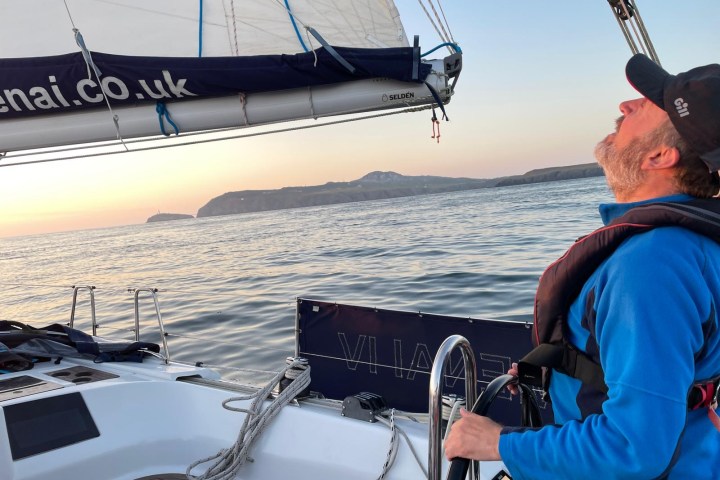 Man steering a sailboat, looking at the sail with an island in the background at sunset.